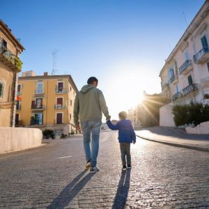 La foto mostra la mano di un genitore che tiene teneramente la mano più piccola di un bambino. Entrambe le mani sono viste da dietro mentre genitore e figlio sembrano camminare insieme verso l'orizzonte, simboleggiando un viaggio condiviso verso il futuro. L'immagine trasmette un senso di guida, supporto e connessione tra genitore e figlio.