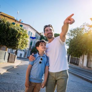 L'immagine mostra un genitore, una figura adulta, che sta in piedi accanto a un bambino, suo figlio. Il genitore tiene in mano un cartello di direzione e sta indicando con l'altra mano in avanti, come per mostrare al figlio la strada da seguire. L'espressione del genitore è seria ma incoraggiante. Il bambino guarda nella direzione indicata con un'espressione attenta e fiduciosa. Lo sfondo è neutro per concentrare l'attenzione sull'interazione tra genitore e figlio.