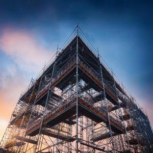 Un'imponente struttura di impalcature metalliche avvolge un edificio in costruzione. Le travi e i tubi d'acciaio si intrecciano in un reticolo geometrico che si estende in altezza, creando livelli multipli di piattaforme di lavoro. L'impalcatura è ancorata alla facciata dell'edificio, fornendo un supporto sicuro per i lavoratori e permettendo l'accesso a tutte le aree della costruzione